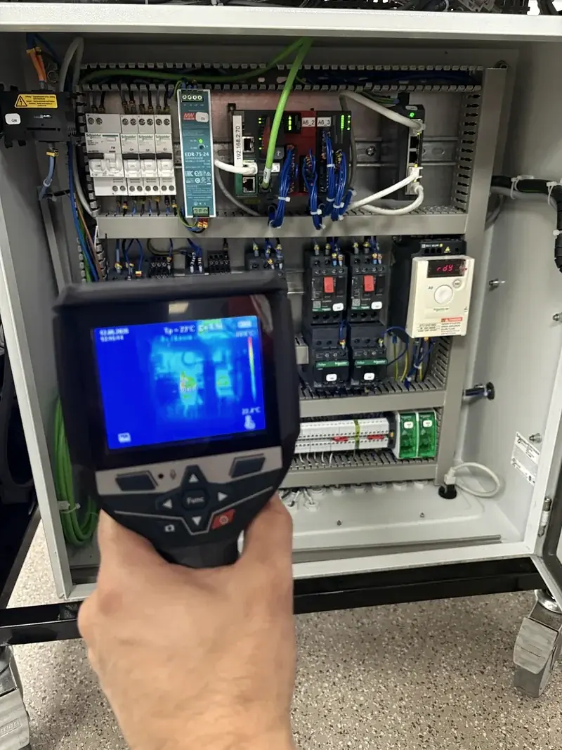 Caméra thermique en main devant un coffret électrique ouvert, image infrarouge visible à l'écran